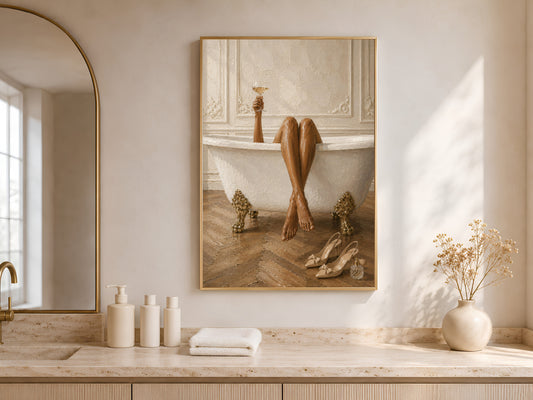 Framed Parisian bath wall art leaning against a warm white wall beside a freestanding soaking tub with neutral ceramics and soft arched window light