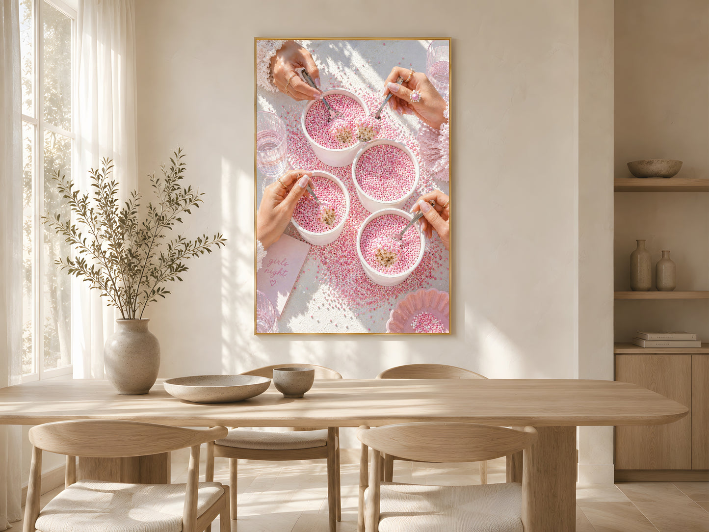 Pink coquette dot cake wall art print in a gold frame above a light oak dining table in a warm Scandinavian dining room with dried botanicals and soft natural light