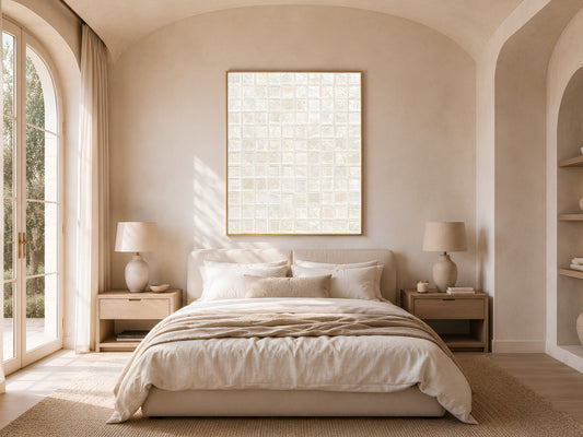 White pearl mosaic tile wall art in a gold frame above a bed with warm linen bedding and a rattan lamp in a bright minimal bedroom with arched windows
