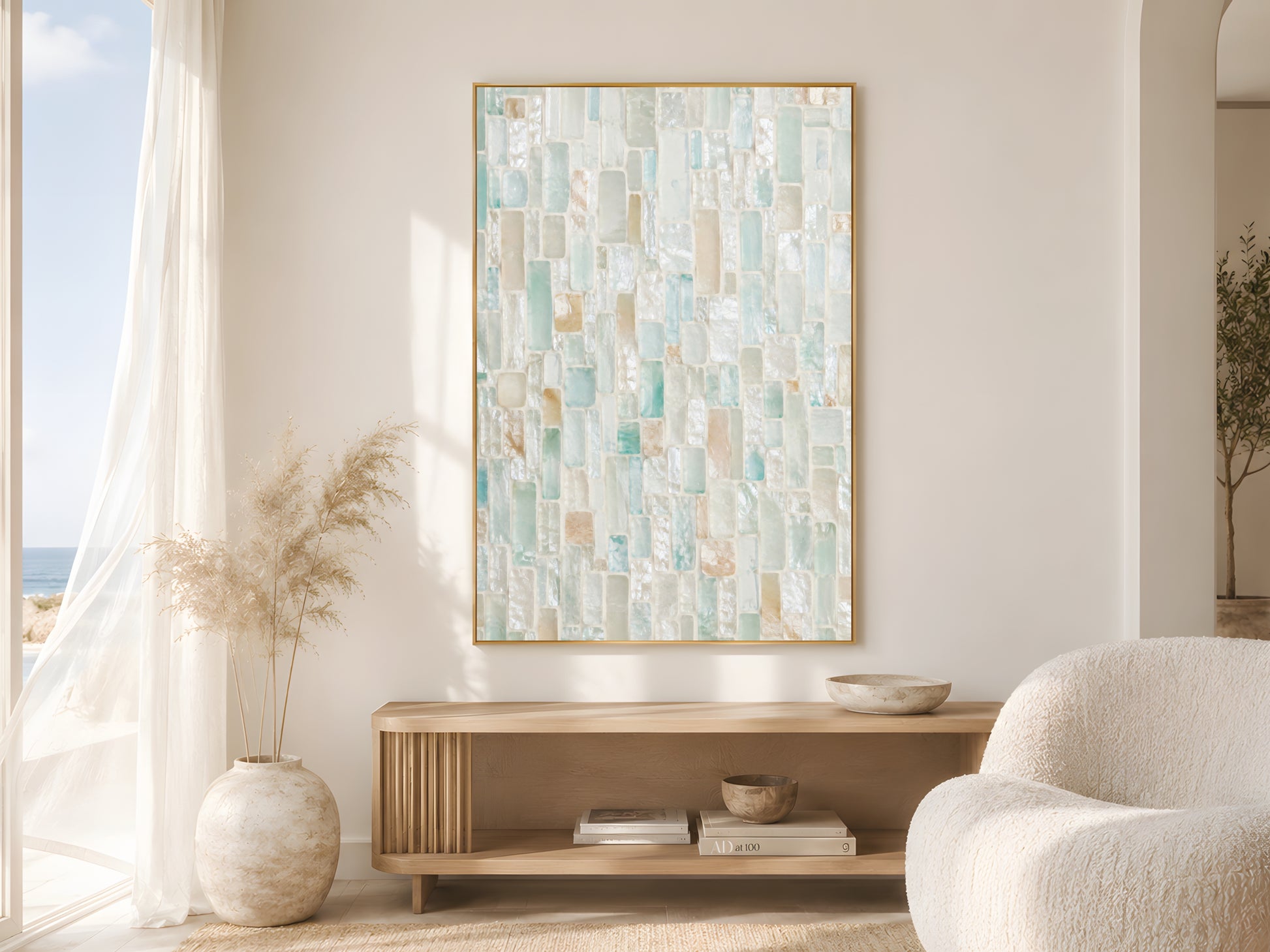 Sea glass mosaic tile print in gold frame beside sheer curtains in bright coastal living room with cream bouclé chair and dried pampas grass
