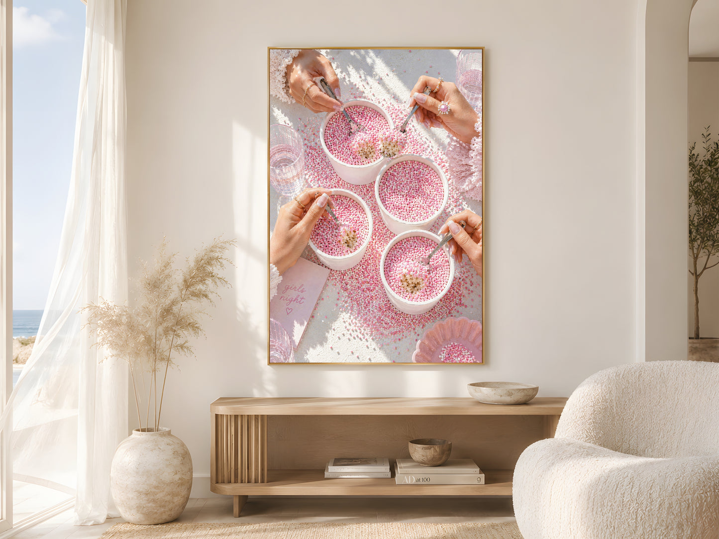 Pink coquette dot cake wall art print in a gold frame above a wooden console in a bright coastal living room with round boucle chair and large windows
