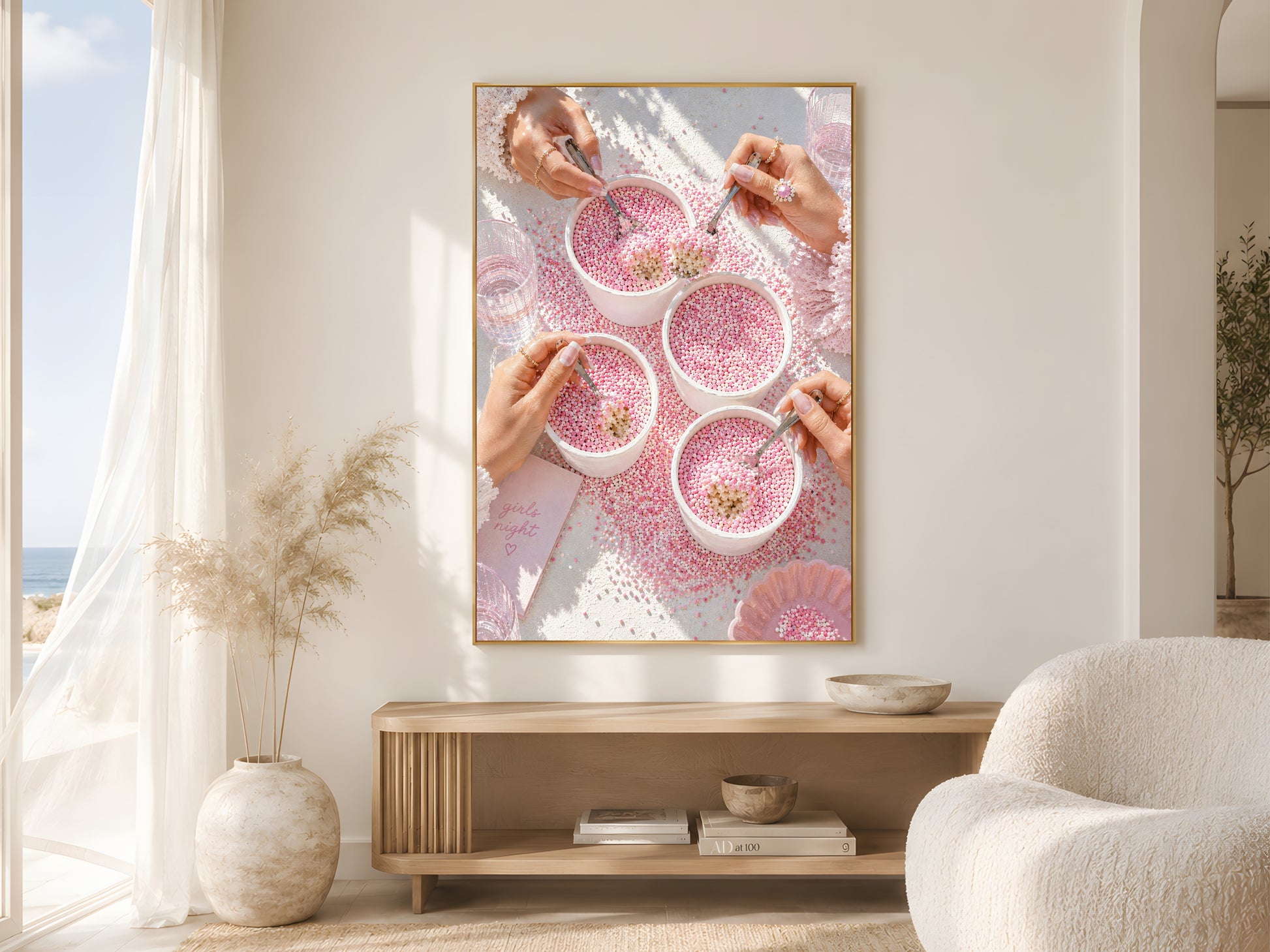 Pink coquette dot cake wall art print in a gold frame above a wooden console in a bright coastal living room with round boucle chair and large windows
