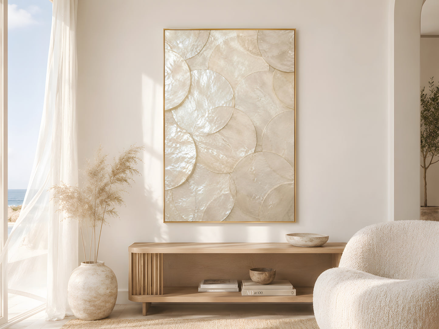 Large capiz shell abstract print in gold frame beside sheer curtains in bright coastal living room with cream bouclé chair and dried pampas grass
