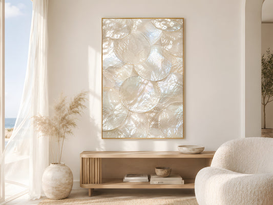 Mother of pearl iridescent shell print in gold frame beside sheer curtains in bright coastal living room with cream bouclé chair and dried pampas grass
