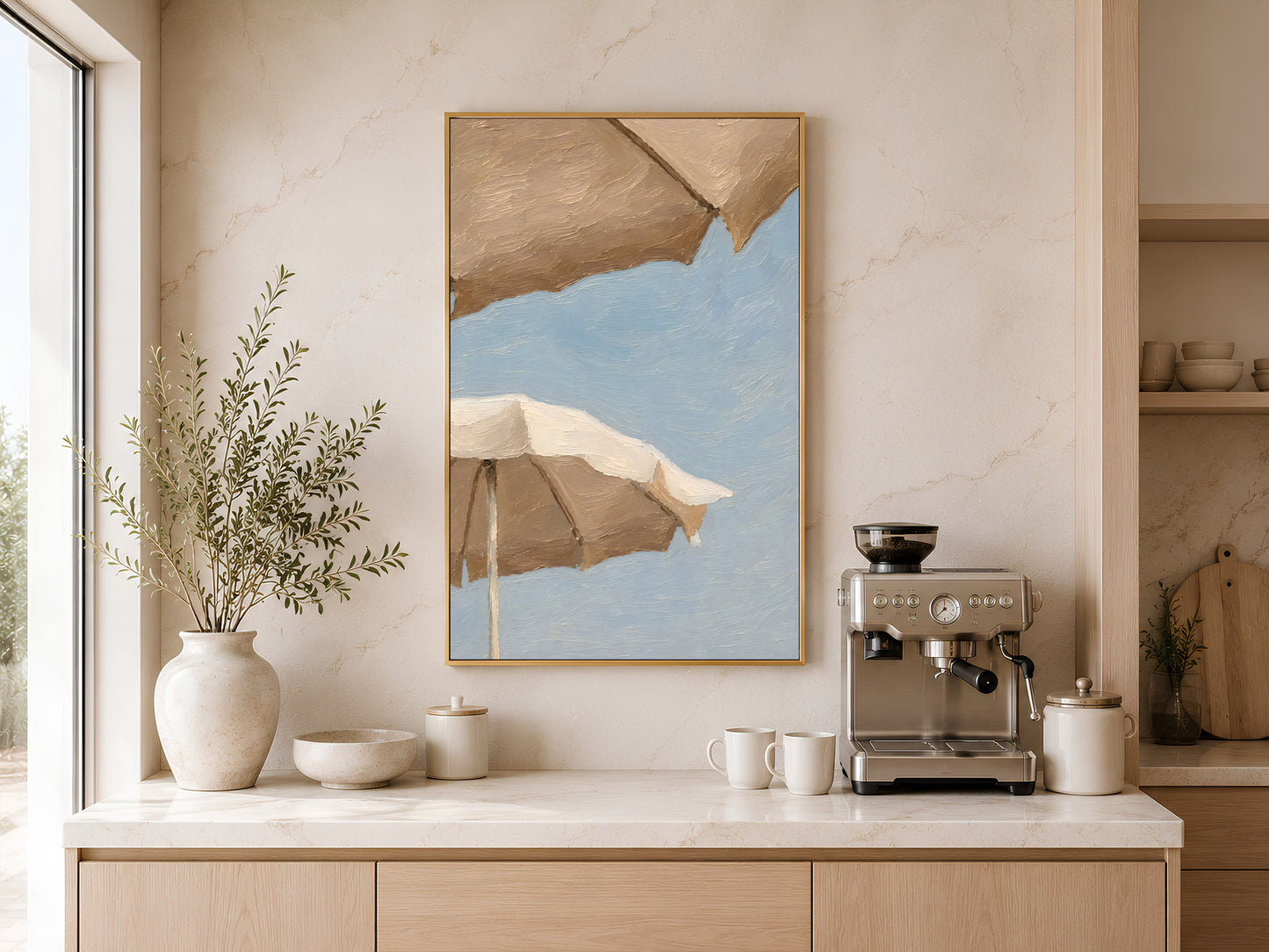 Beach umbrella wall art in a gold frame displayed above a coffee station in a warm minimalist kitchen with cream cabinetry and dried greenery
