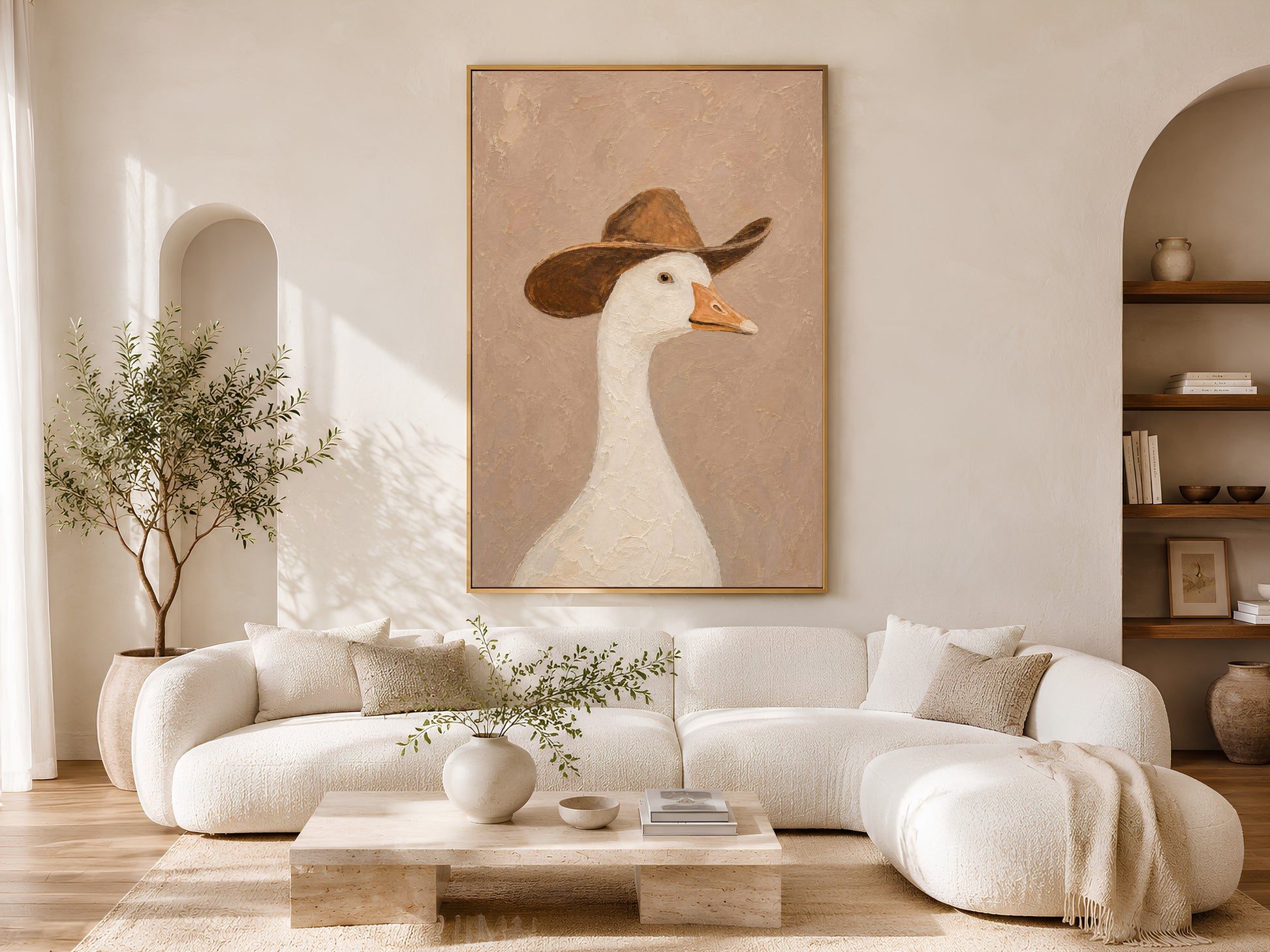 Cowboy goose wall art in a gold frame above a cream sofa in a bright neutral living room with an olive tree, warm wood flooring, and soft arched architectural details


