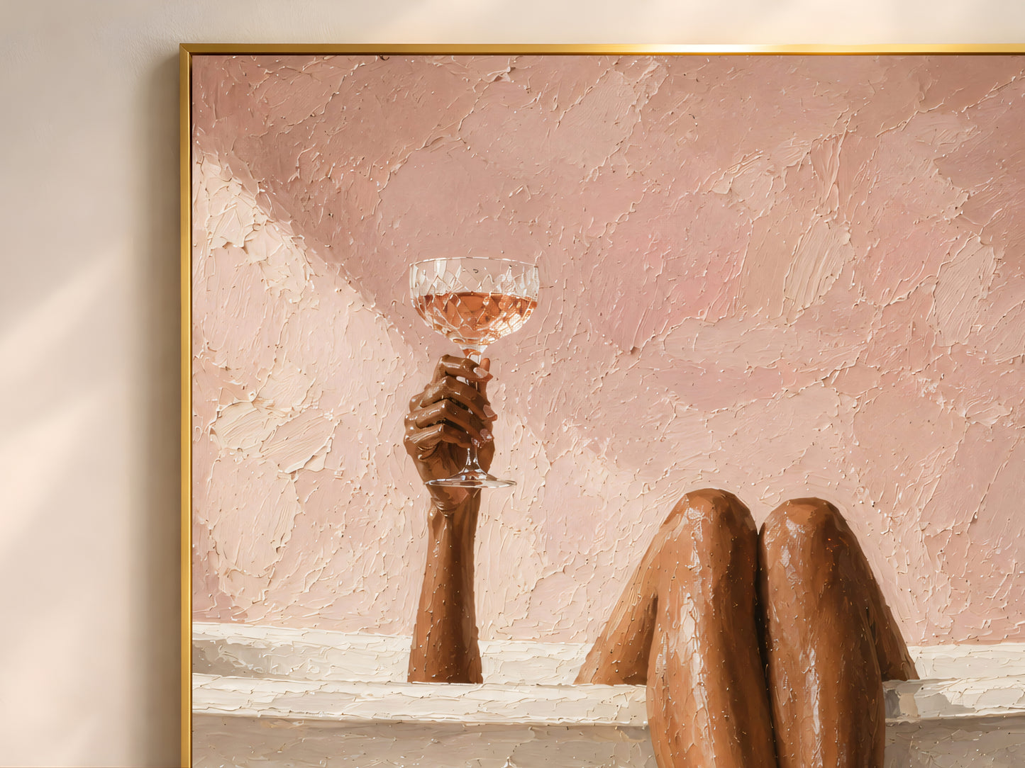 Close-up of the blush bath art print in a gold frame showing bronzed legs over a cream tub and a hand raising a crystal rosé coupe against a warm dusty pink impasto-textured wall