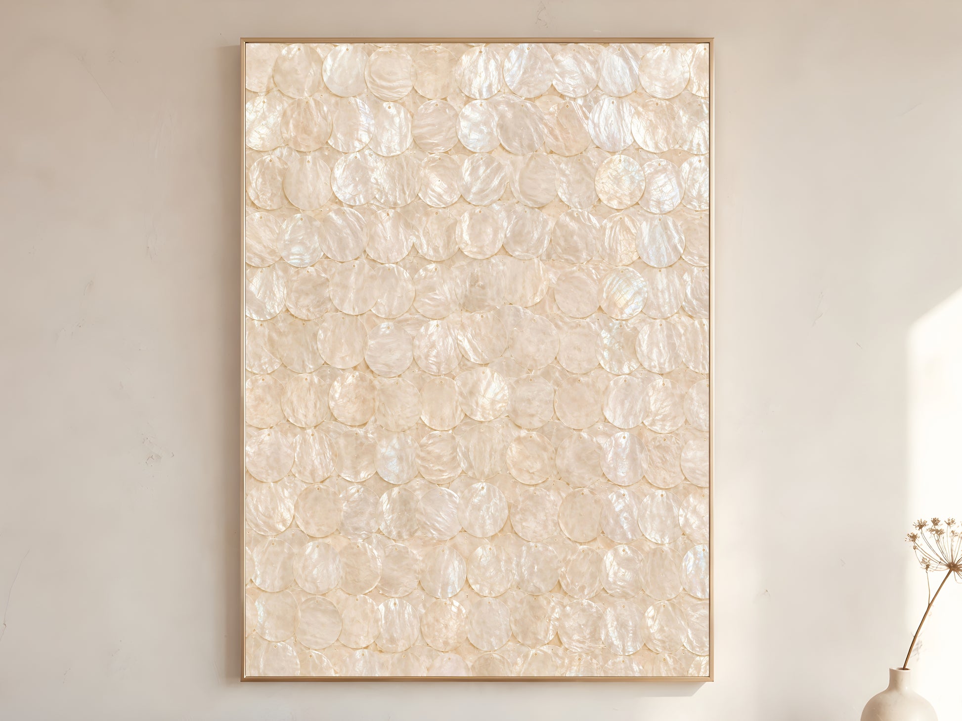 Capiz shell wall art print in a gold frame leaning against a warm white wall with dried botanicals, showing the full pattern of overlapping iridescent ivory and cream shell discs with soft pearlescent texture

