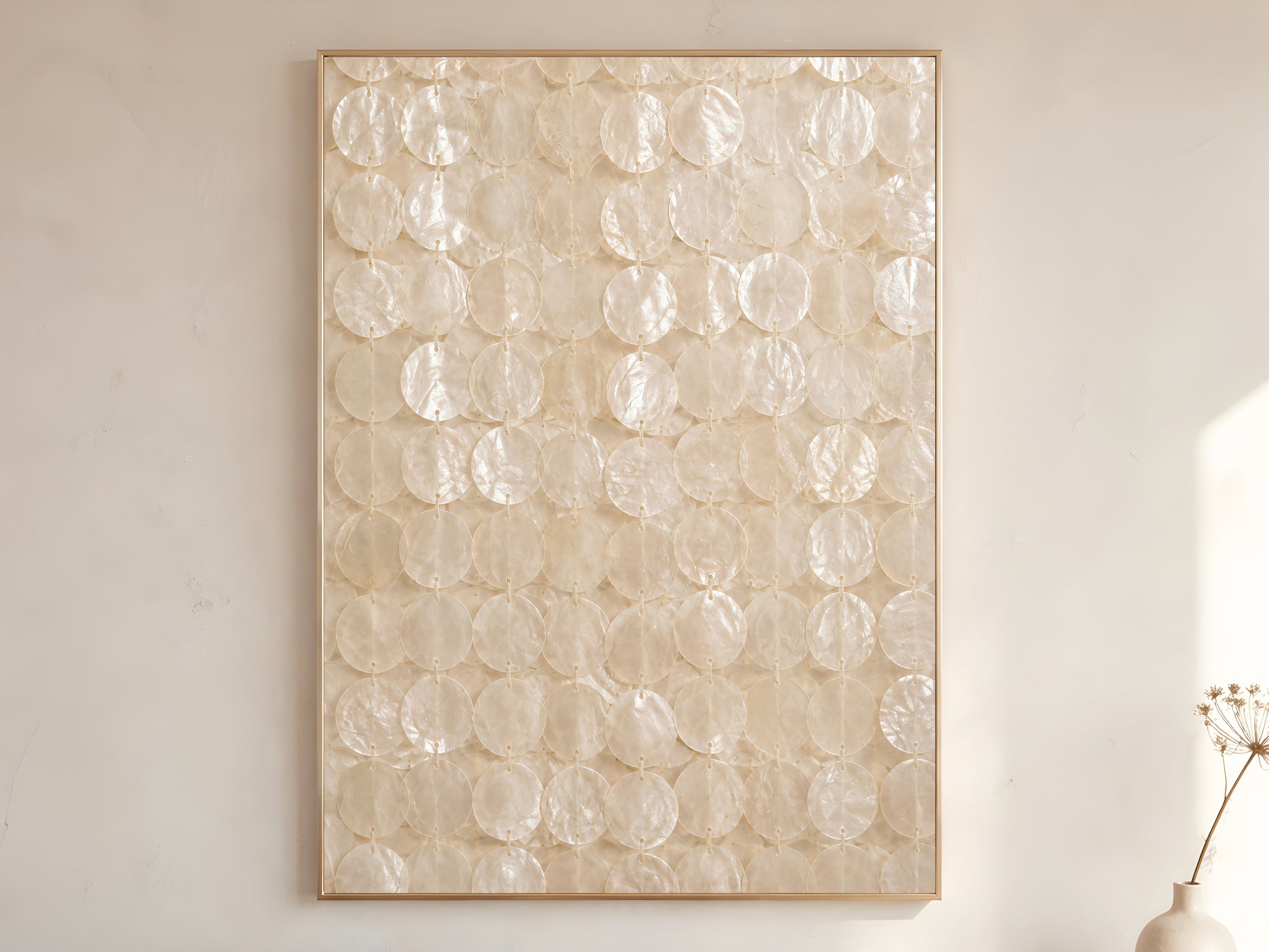 Capiz shell wall art print in a gold frame leaning against a warm white wall with dried botanicals, showing the full pattern of translucent ivory shell discs strung in vertical rows with soft iridescent pearlescent texture
