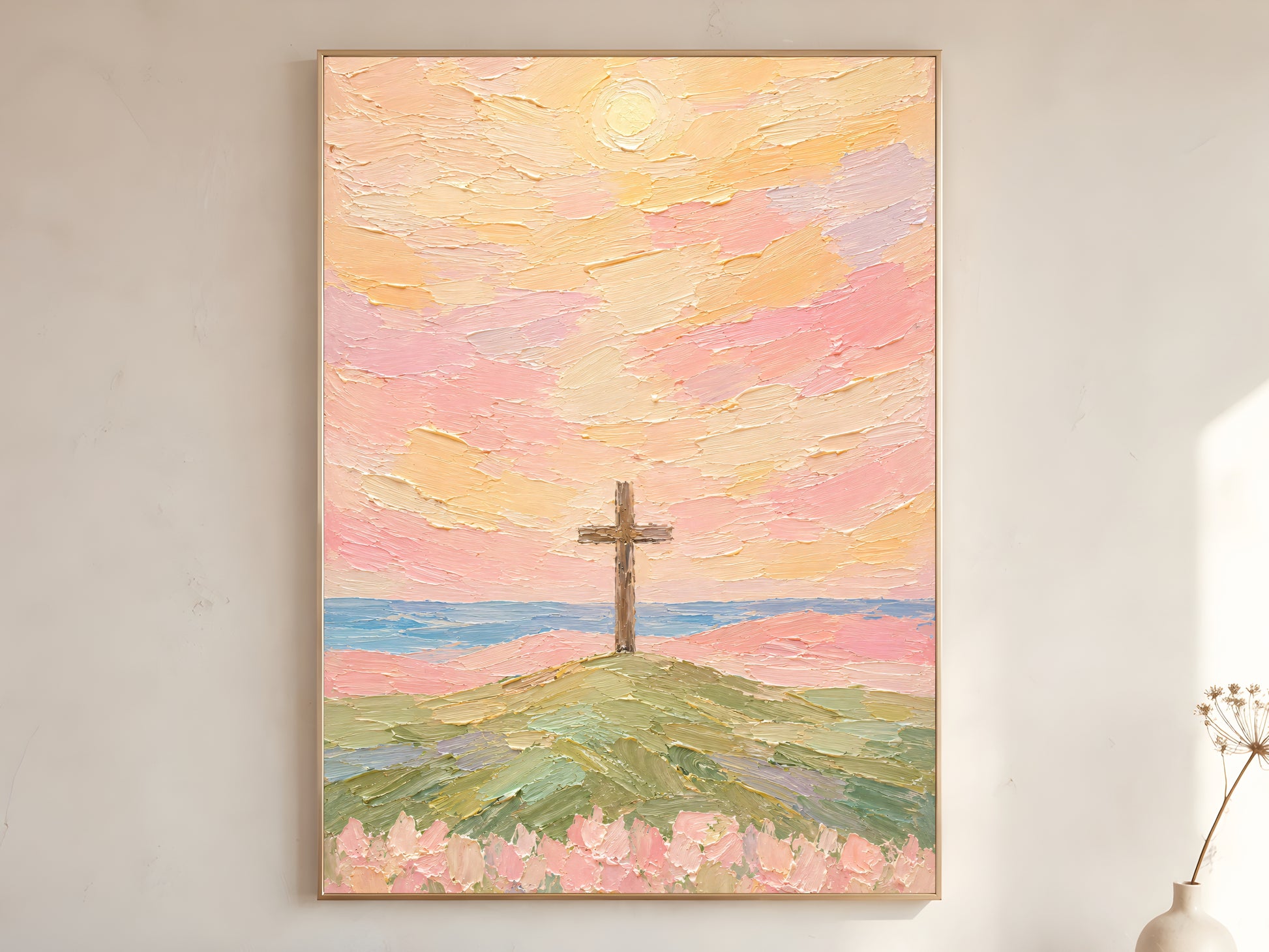 Cross wall art print framed and hanging on a neutral wall with a vase and dried branches nearby, pastel sunset sky over hill and ocean.