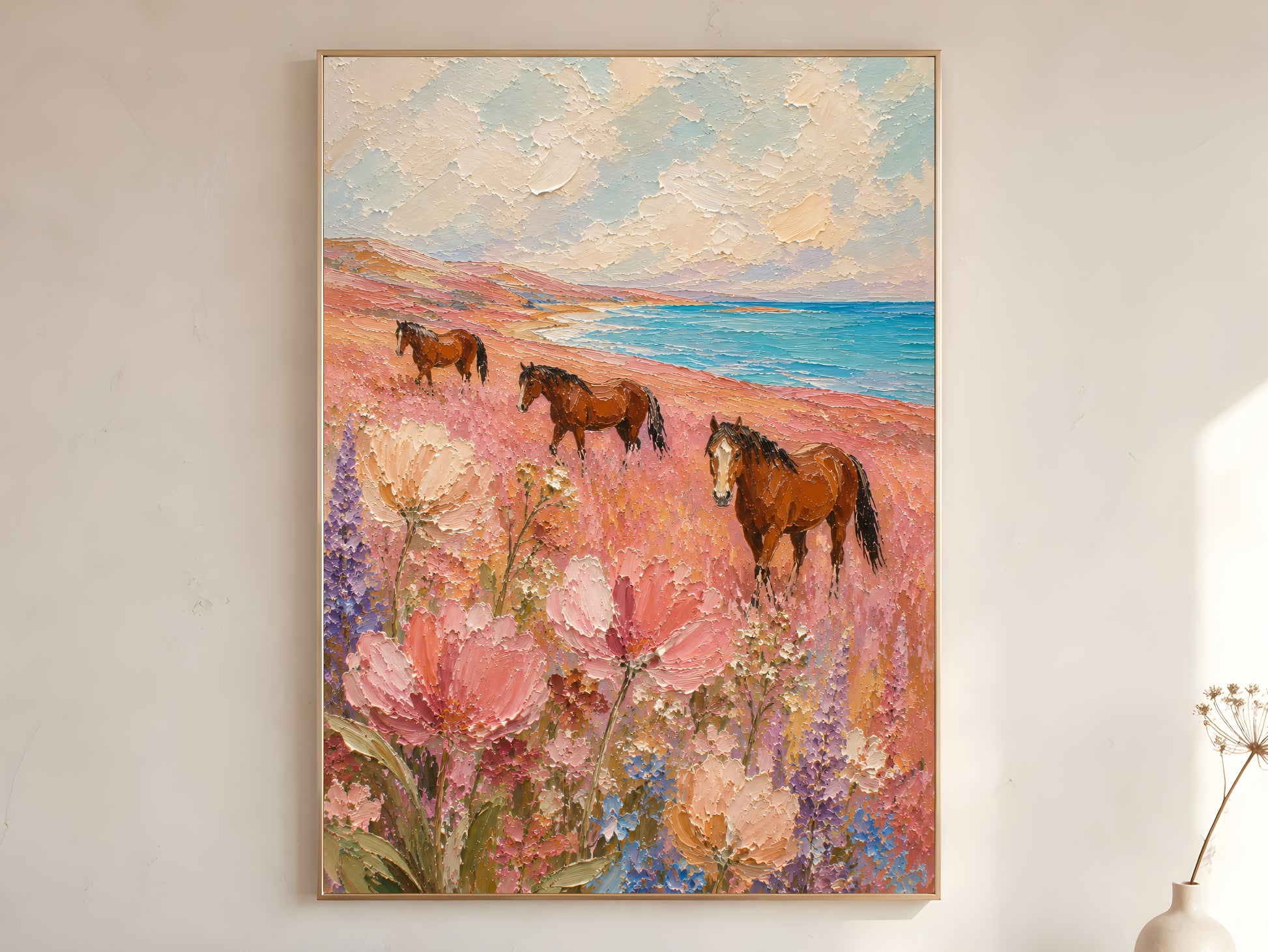 Three chestnut horses in a pink wildflower coastal meadow impasto oil painting print in a gold frame leaning against a warm white wall with dried botanicals, showing the full painting with turquoise ocean, creamy sky, and large pink cosmos in the foreground

