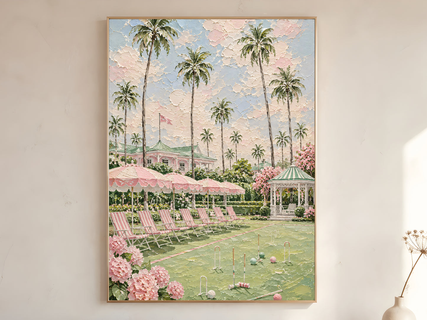 Pink country club croquet lawn with tall palm trees, striped parasols, white gazebo, and pink hydrangeas impasto oil painting print in a thin natural wood frame, leaning against a warm white wall with a dried wildflower stem
