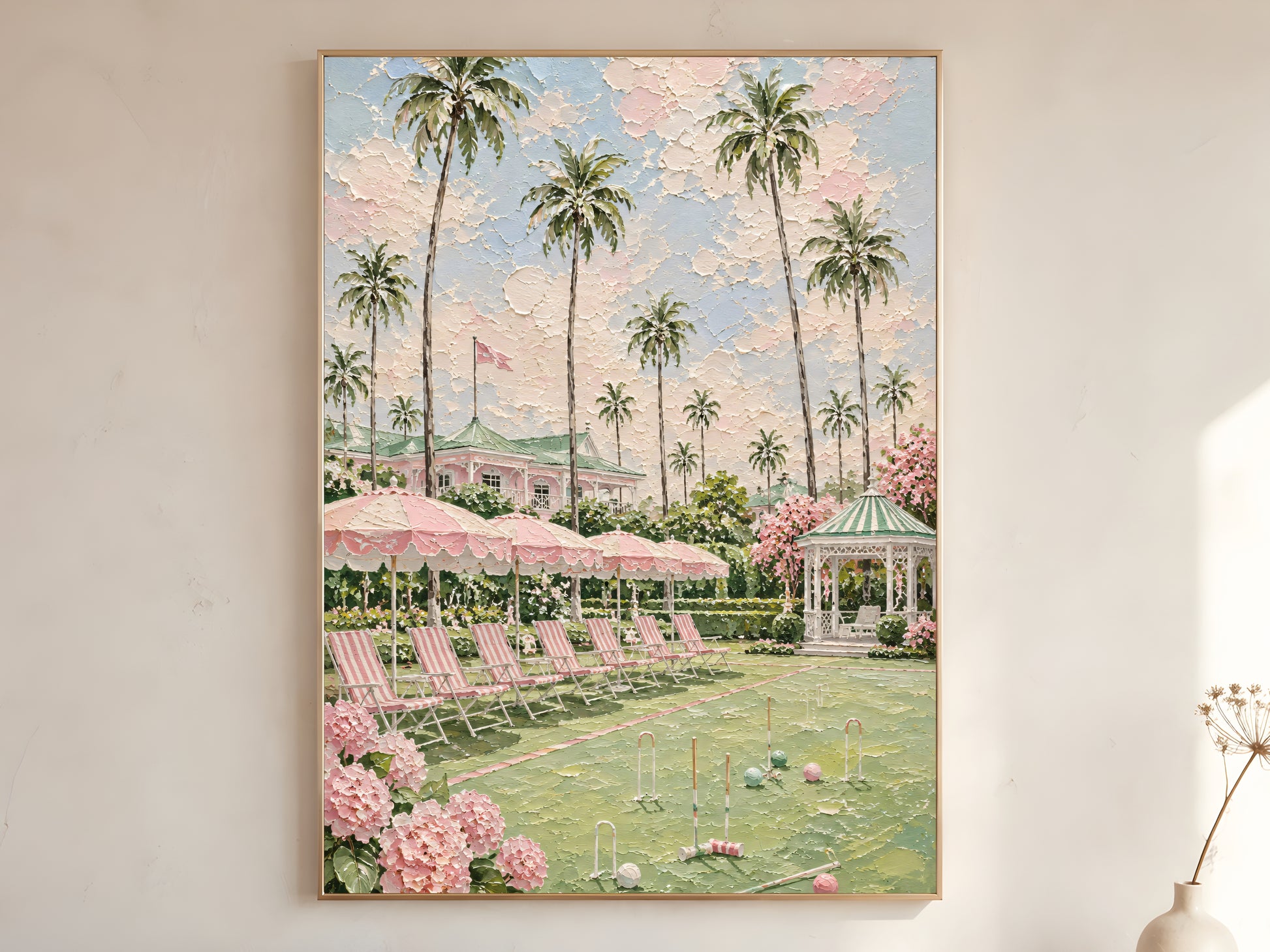 Pink country club croquet lawn with tall palm trees, striped parasols, white gazebo, and pink hydrangeas impasto oil painting print in a thin natural wood frame, leaning against a warm white wall with a dried wildflower stem
