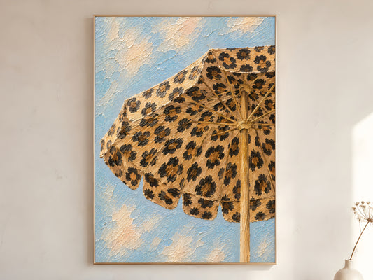 Leopard print beach umbrella impasto wall art print against a blue sky in a gold frame leaning against a warm white wall with dried stems
