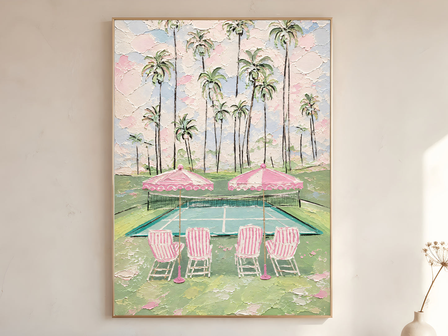 Pink tennis court with tall palm trees, striped chairs, and pink parasols impasto oil painting print in a thin natural wood frame, leaning against a warm white wall with a dried wildflower stem
