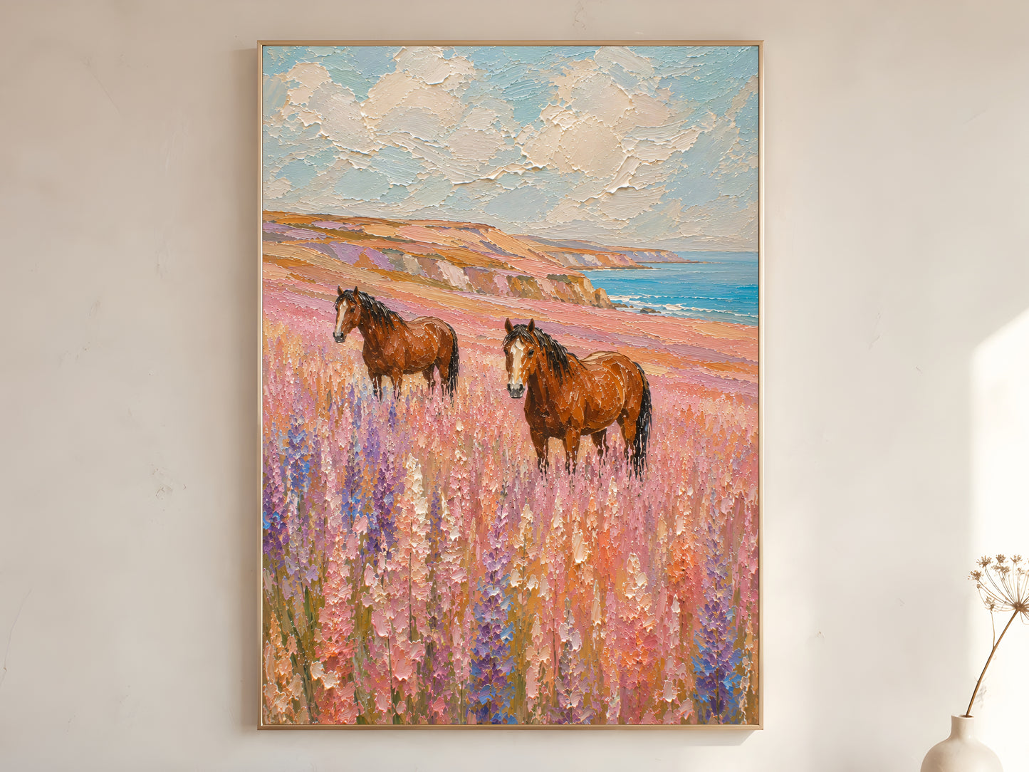 Two chestnut horses standing in a lavender and pink wildflower coastal meadow impasto oil painting print in a gold frame leaning against a warm white wall with dried botanicals, showing the full painting with turquoise ocean, amber cliffs, and creamy impasto sky