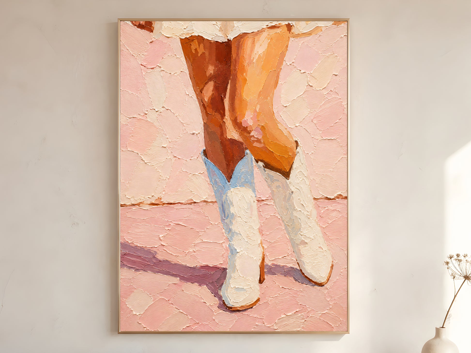 Large framed pink cowgirl boots impasto wall art print showing white cowboy boots and bronzed legs on blush pink background displayed on warm white wall with dried flower stem
