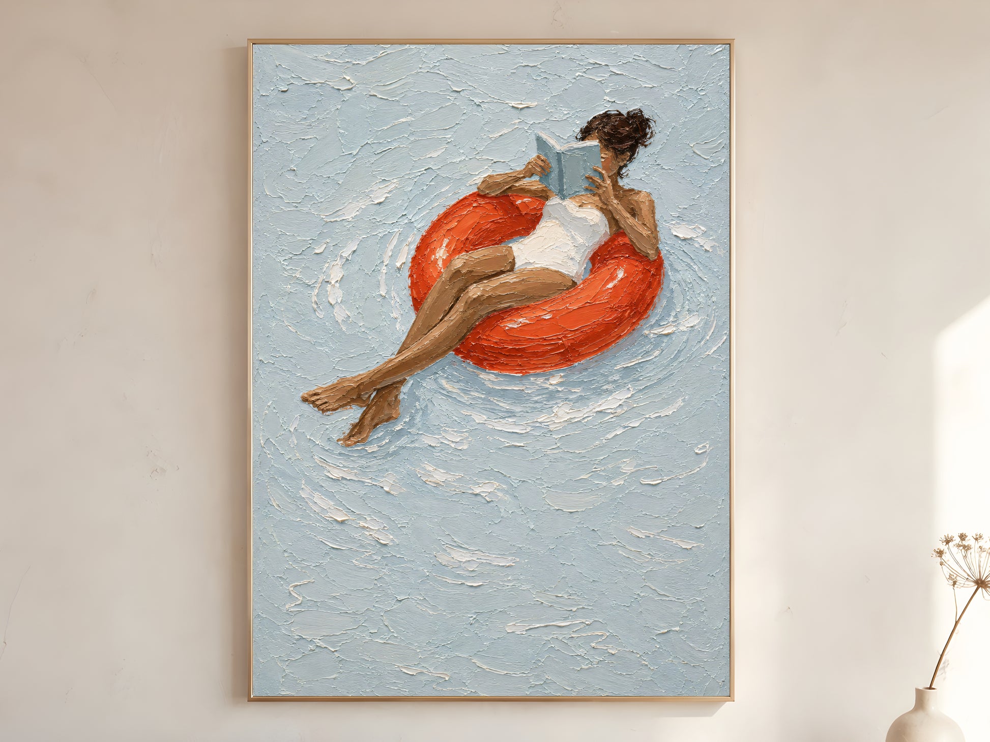 Woman reading on red pool float impasto oil painting print in a thin natural wood frame, leaning against a warm white wall with a dried wildflower stem

