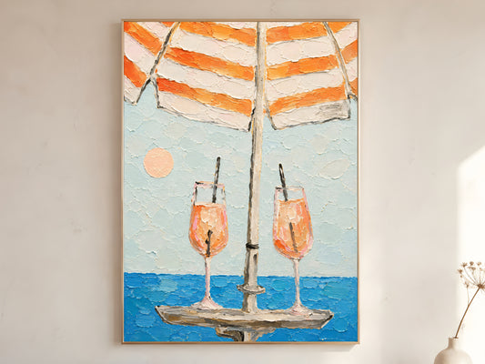 Large framed aperol spritz beach umbrella impasto wall art print showing two cocktail glasses under orange striped parasol with teal ocean displayed on warm white wall with dried flower stem
