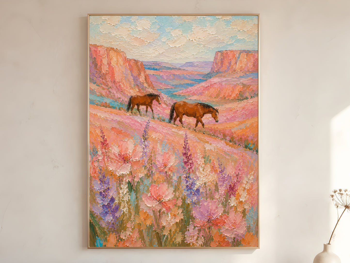 Two chestnut horses walking through a pink and lavender wildflower canyon impasto oil painting print in a gold frame leaning against a warm white wall with dried botanicals, showing the full painting with terracotta canyon walls, turquoise river valley, and creamy impasto sky

