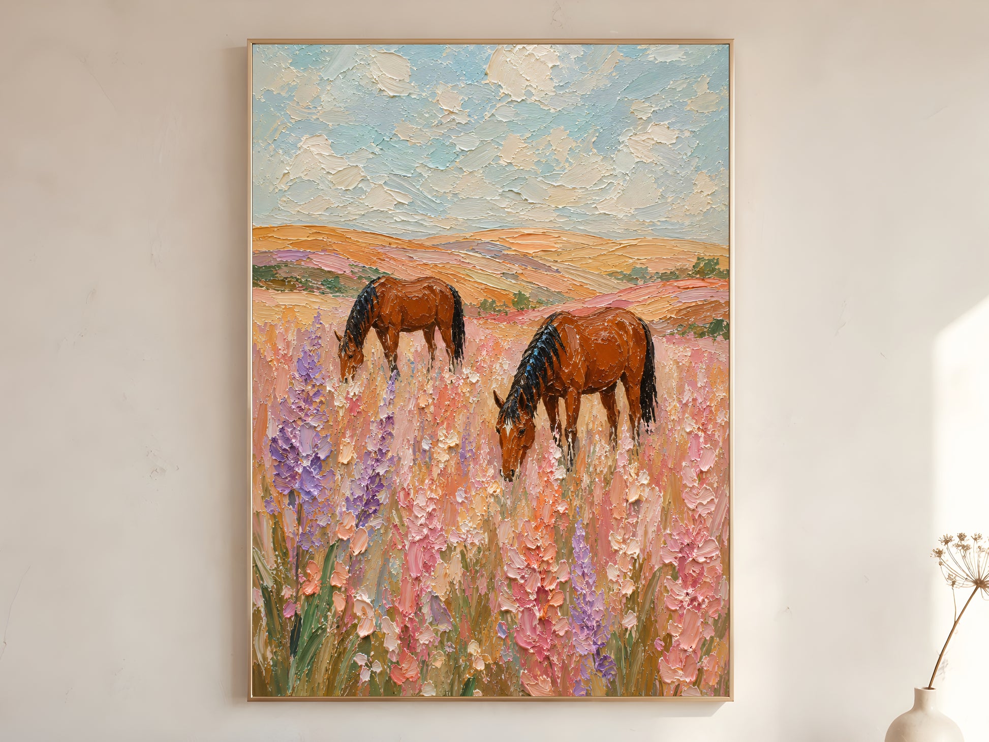 Two chestnut horses grazing in a pink and lavender wildflower field impasto oil painting print in a gold frame leaning against a warm white wall with dried botanicals, showing the full painting with chalky sky and rolling golden hills

