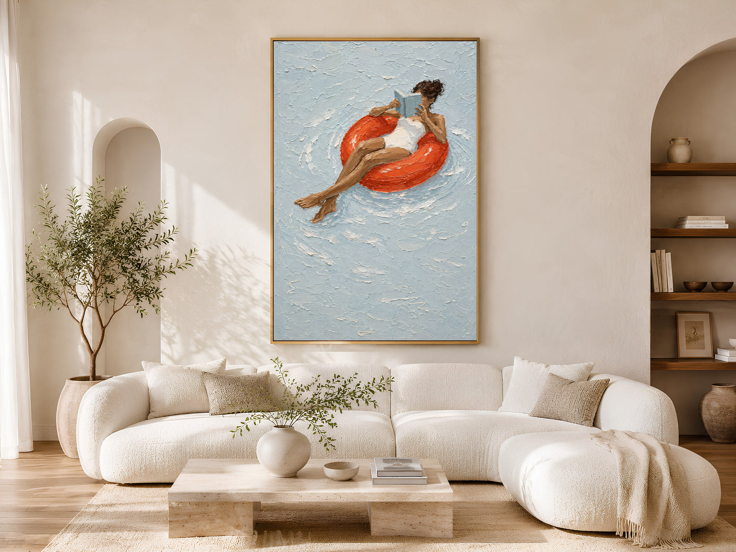 Gold-framed pool float reading girl wall art above a cream curved sofa in a bright minimalist living room with arched wall detail and olive tree
