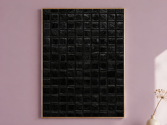 Black impasto mosaic tile wall art in a thin gold frame leaning against a dusty mauve wall with a dried allium stem in a cream vase beside it

