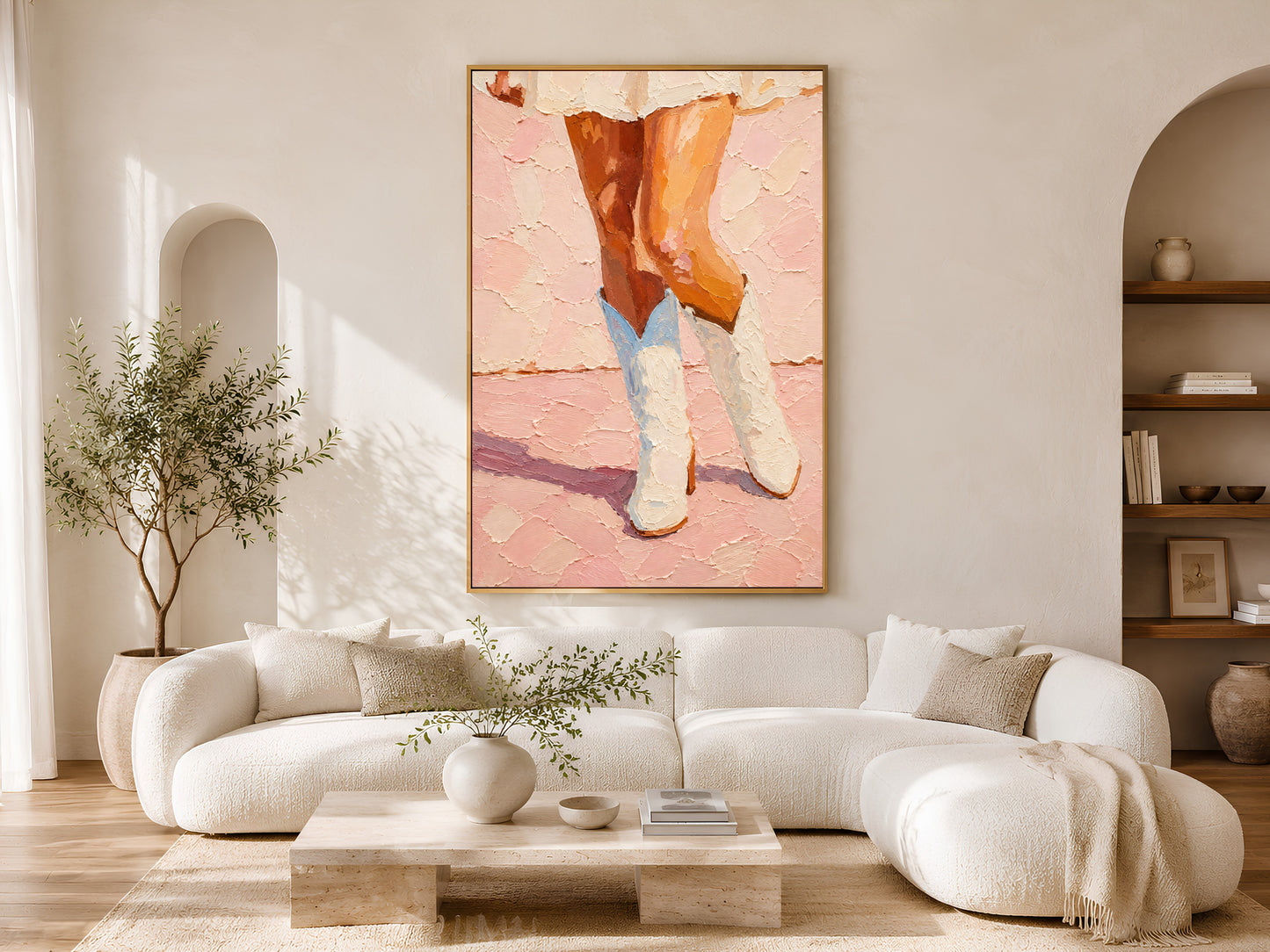 Framed pink cowgirl boots impasto print on white wall in a bright Scandinavian living room with curved cream sofa, round coffee table, and olive tree
