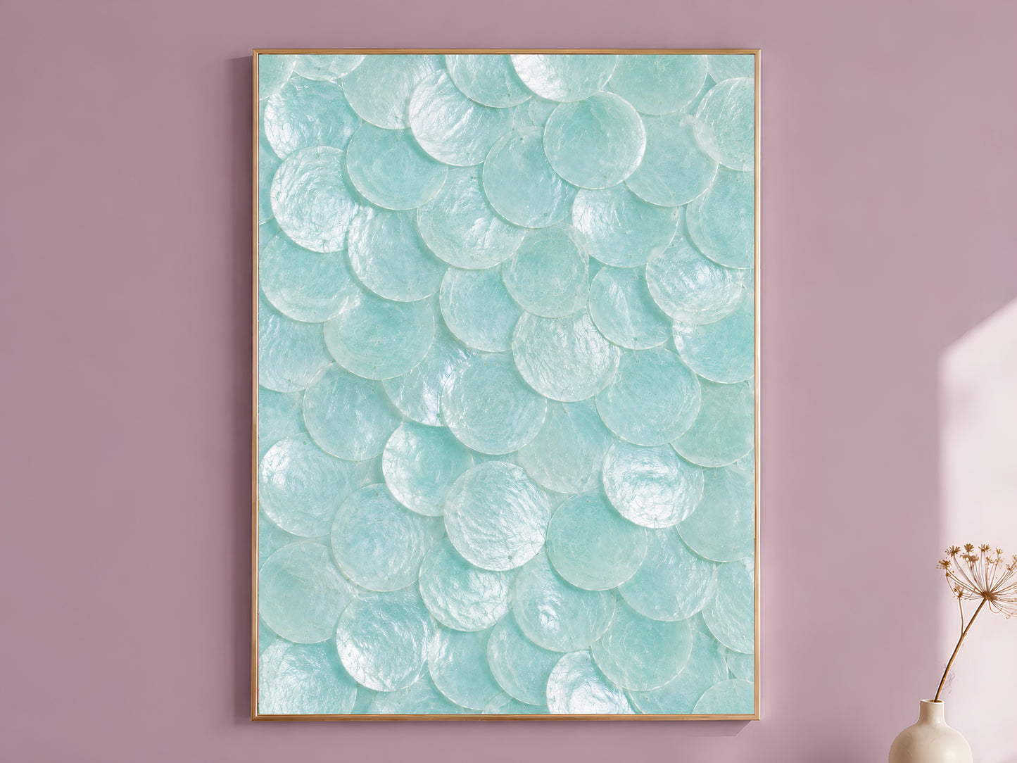 Seafoam capiz shell wall art in a thin gold frame, hung on a mauve wall with a vase of dried flowers.
