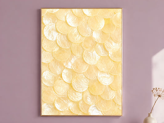 Capiz shell wall art in butter yellow and ivory, framed in thin gold, hung on a dusty mauve wall beside a vase of dried flowers

