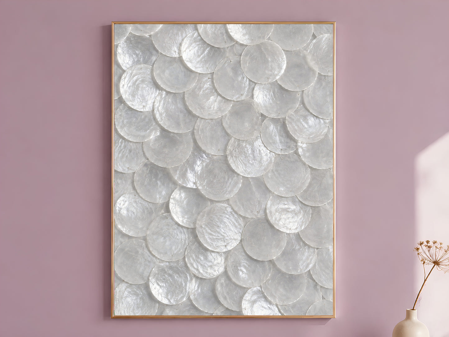 White capiz shell wall art in a thin gold frame, hung on a mauve wall with a vase of dried flowers.
