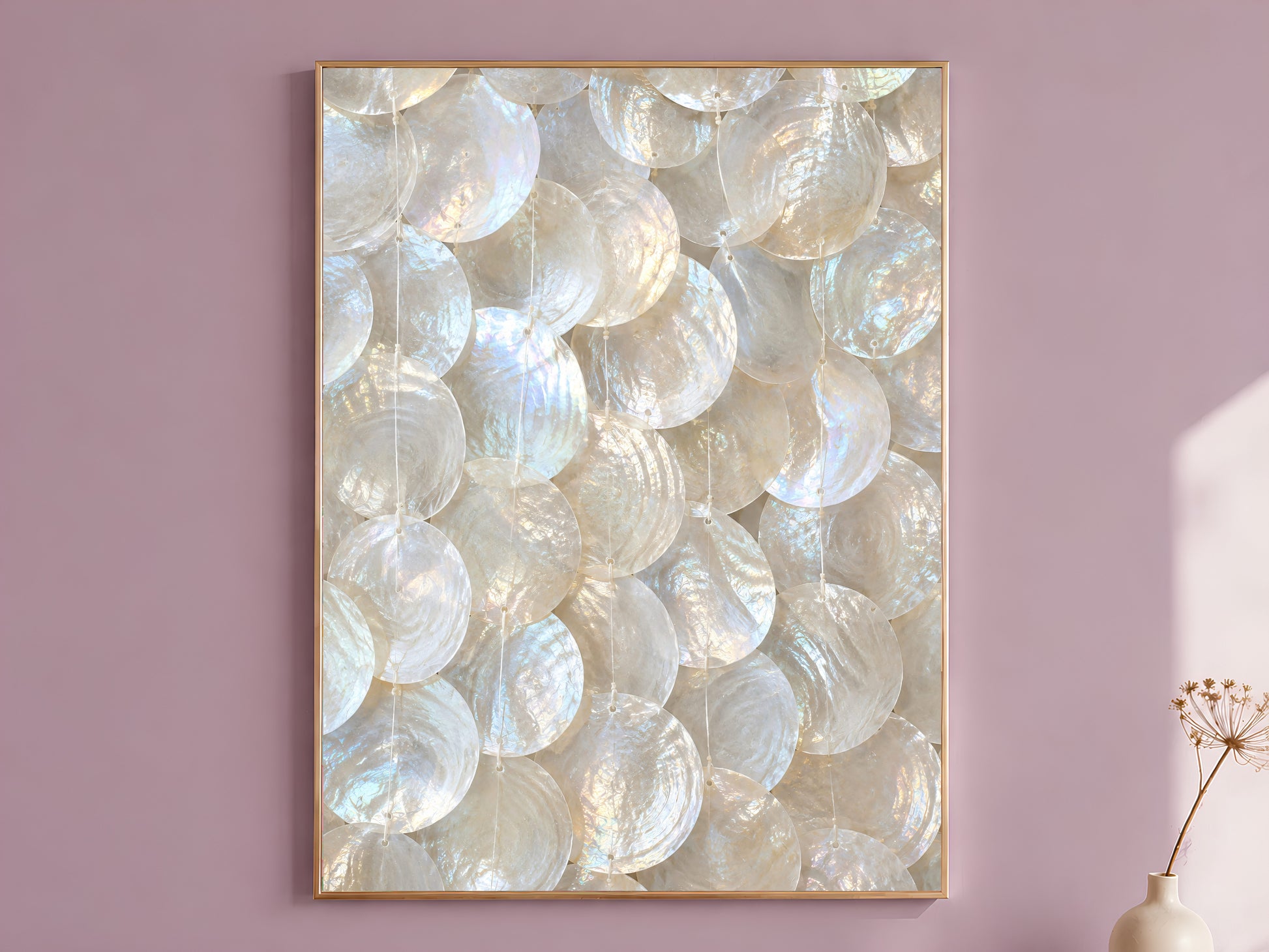 Capiz shell wall art print in a gold frame hanging on a dusty mauve wall beside a vase of dried flowers.