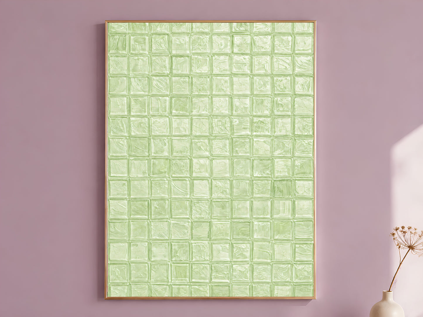 Sage green impasto mosaic tile wall art in a thin gold frame leaning against a dusty mauve wall with a dried allium stem in a cream vase beside it

