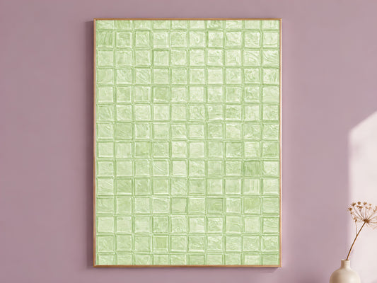 Sage green impasto mosaic tile wall art in a thin gold frame leaning against a dusty mauve wall with a dried allium stem in a cream vase beside it
