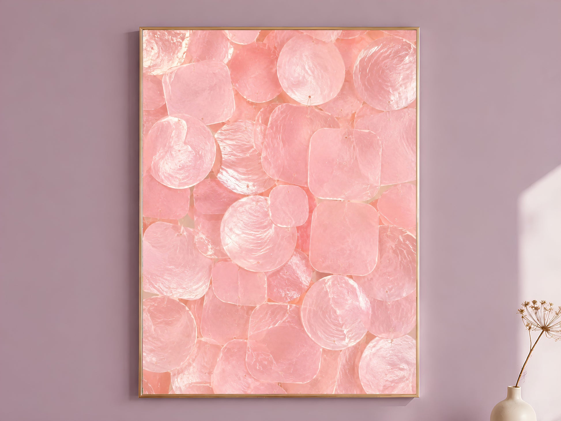 Pink capiz shell wall art print in a thin gold frame, hung on a mauve wall with a vase of dried flowers.
