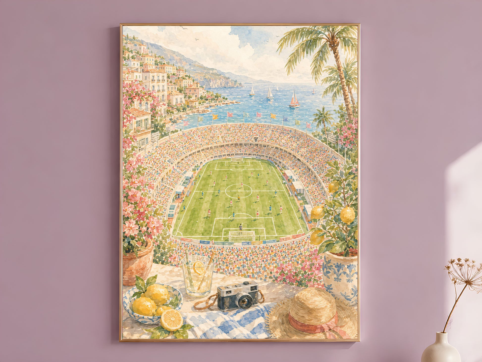 Framed coastal football stadium wall art print in a thin gold frame, hanging on a dusty mauve wall above a wooden bench with a dried flower stem.
