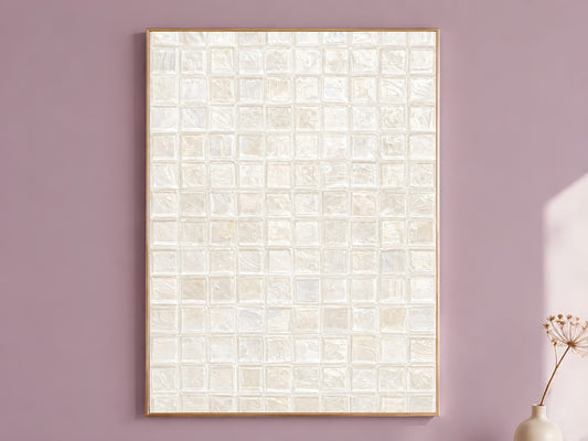 White pearl impasto mosaic tile wall art in a thin gold frame leaning against a dusty mauve wall with a dried allium stem in a cream vase beside it
