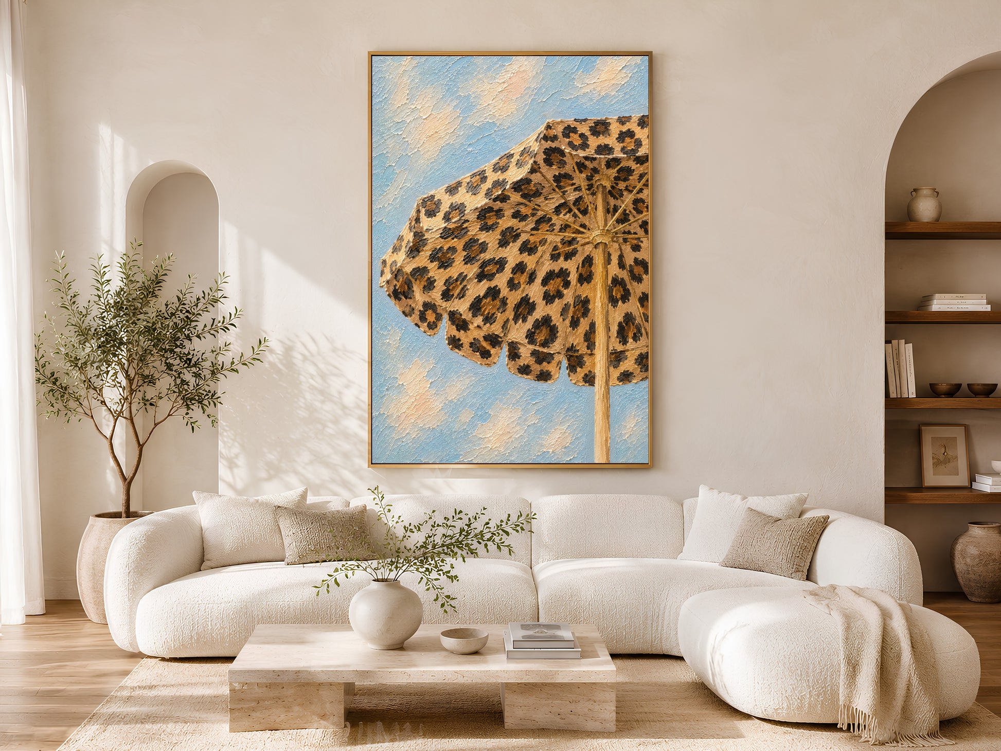 Framed leopard print beach umbrella wall art in a warm neutral living room with white curved sofa, round coffee table, olive tree and arched alcove
