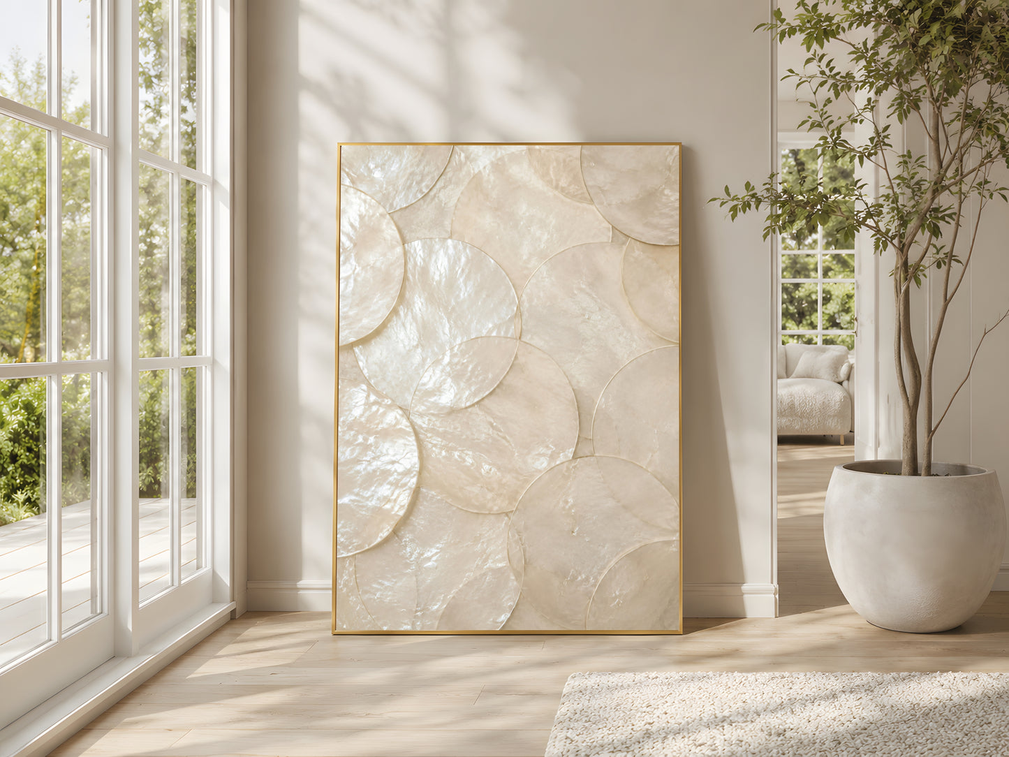 Large capiz shell pearlescent wall art in gold frame beside floor-to-ceiling windows in bright Mediterranean living room with indoor olive tree
