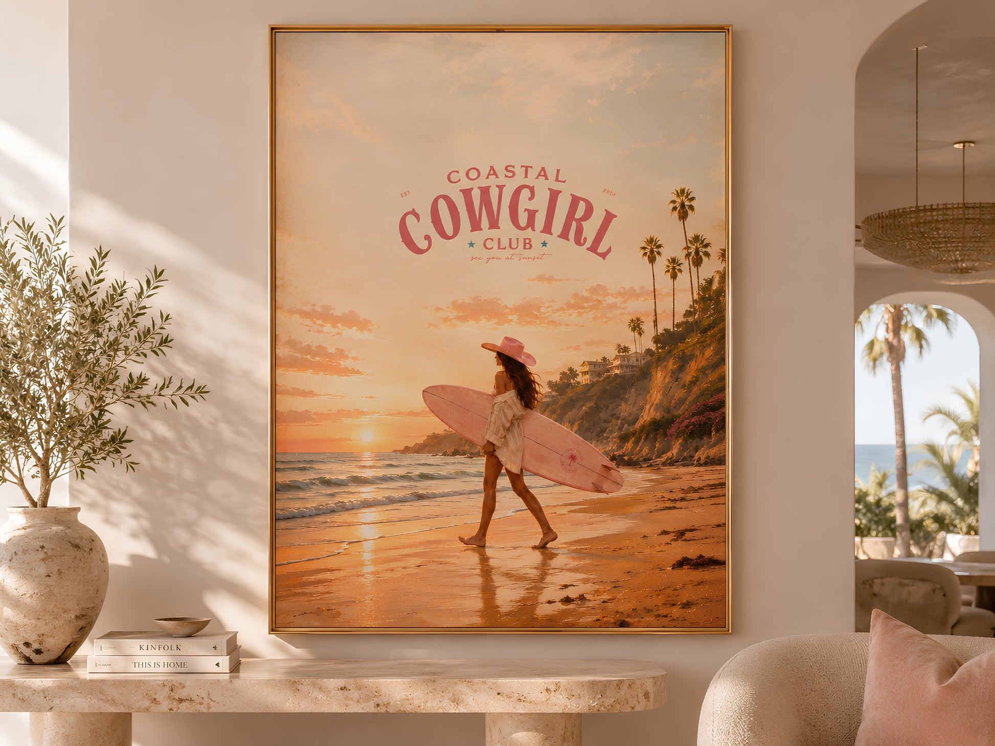 Coastal Cowgirl Club surfer girl sunset print in a gold frame displayed on a wooden console table in a warm neutral living room with olive tree and arched doorway

