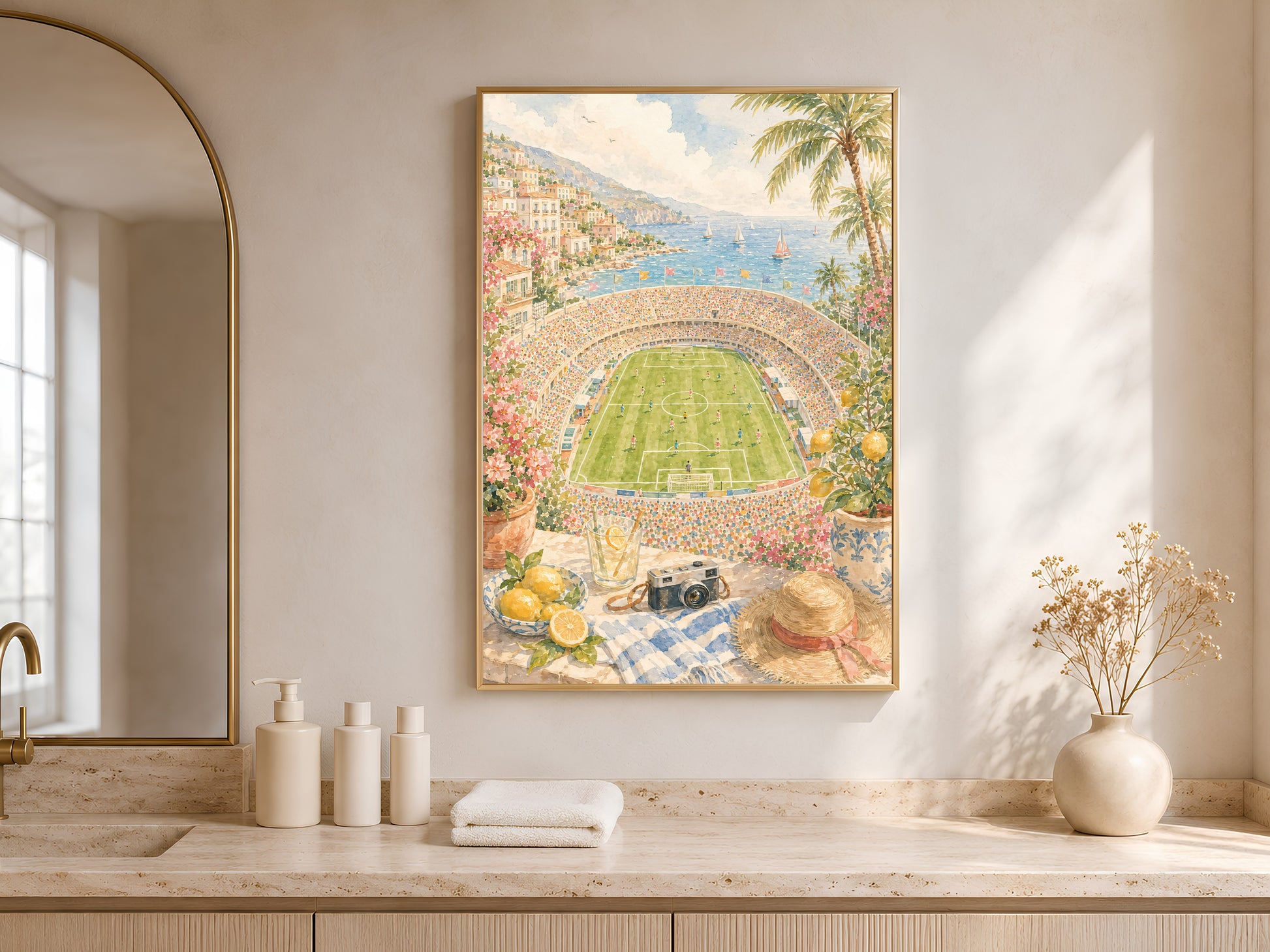 Bathroom vanity styled with folded towels and glass bottles beside a sunlit window, framed coastal football stadium print hanging above the counter.

