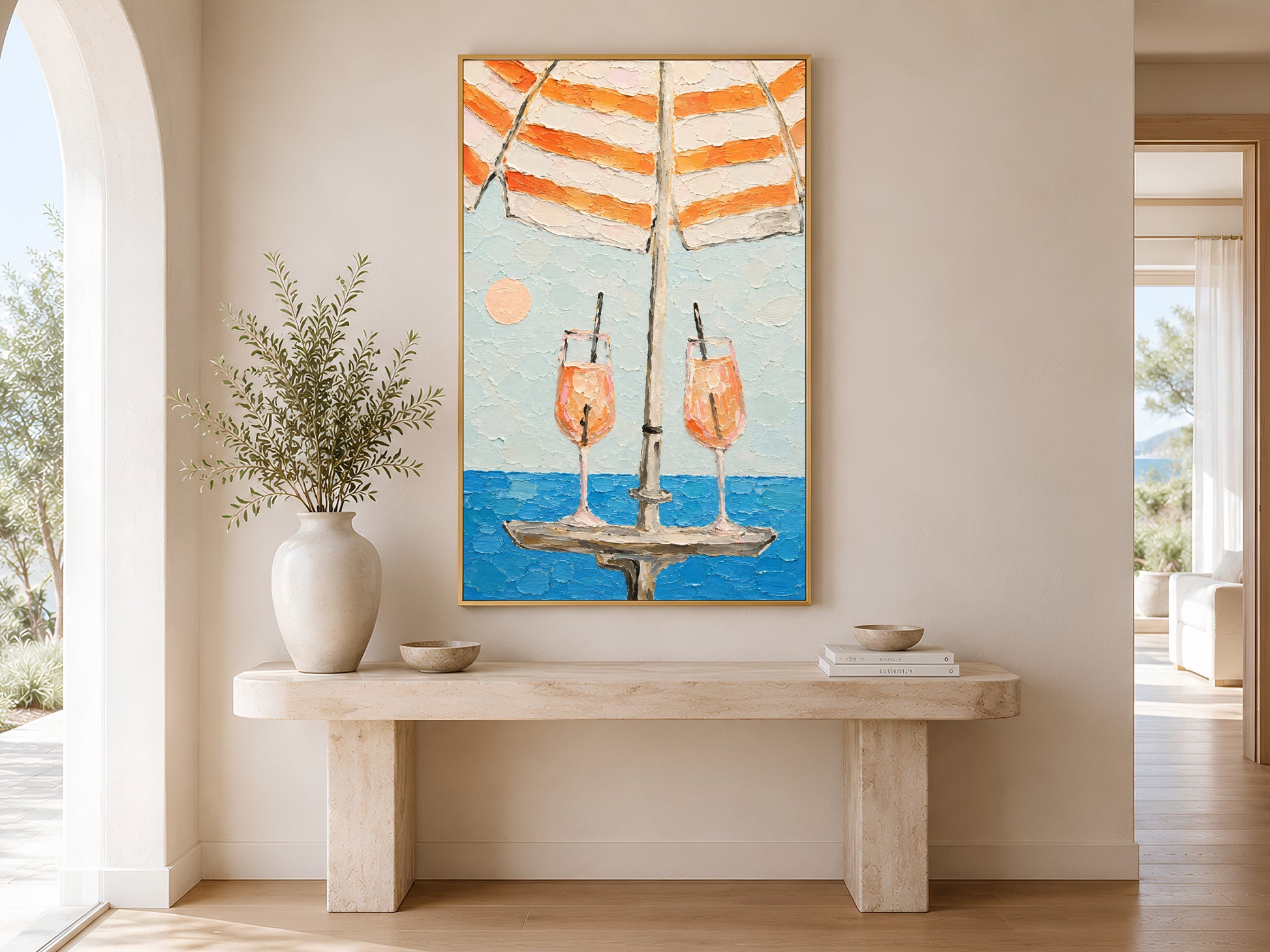 Framed aperol spritz beach umbrella wall art displayed above a wooden console table with large white ceramic vase in a minimal neutral entryway
